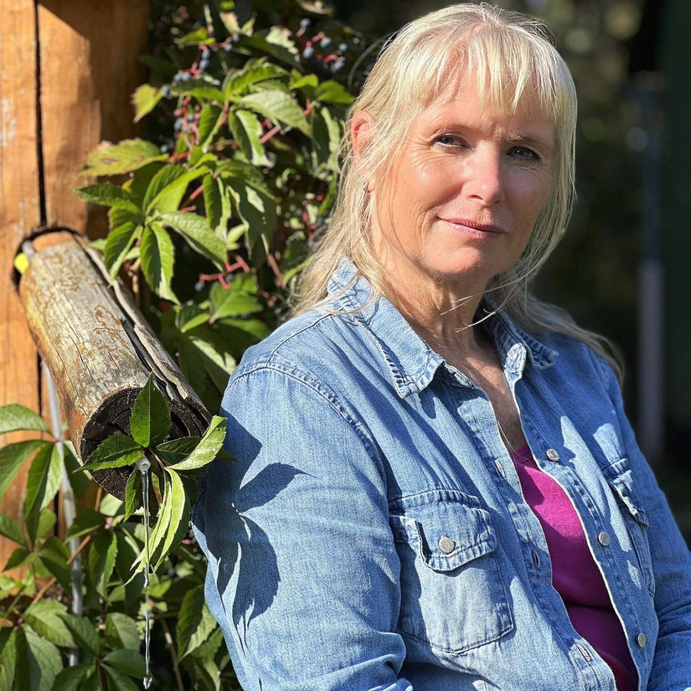 Frau mit blonden Haaren, Jeansjacke und pinkem Oberteil, lächelt. Im Hintergrund eine grüne Pflanze.