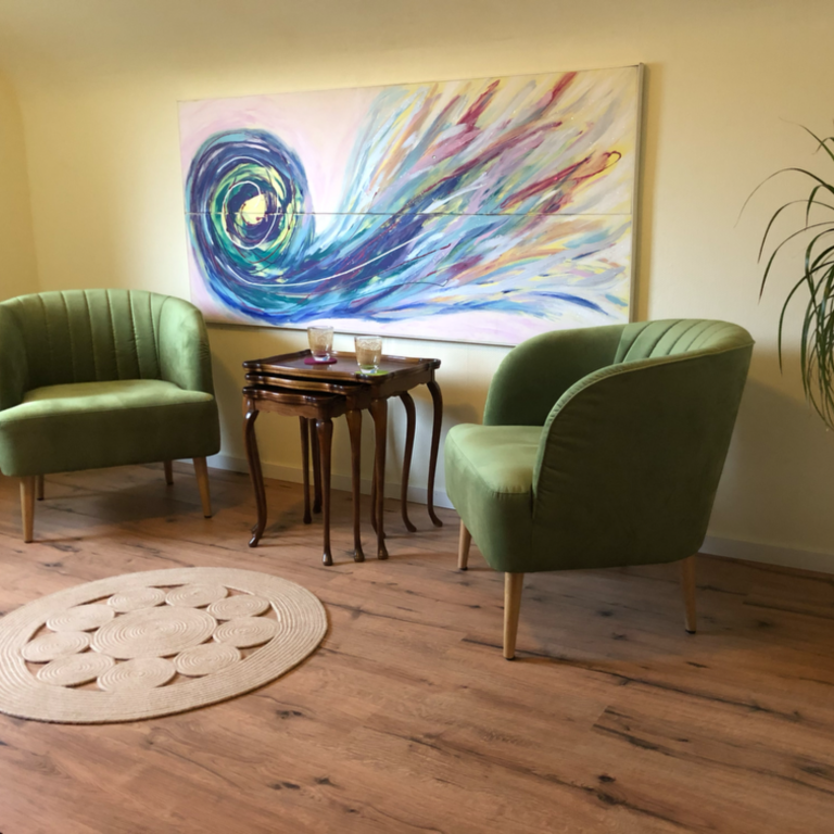 Zwei grüne Sessel stehen an einem kleinen Tisch, vor einem bunten Wandbild und einem Teppich.