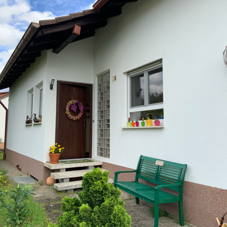 Eingang eines Hauses mit grüner Bank, blühenden Pflanzen und bunter Fensterdekoration.