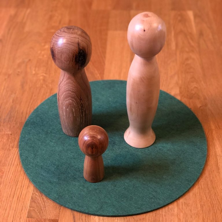 Drei Holzfiguren unterschiedlicher Größe auf einem grünen Filzuntergrund symbolisieren eine Familie.