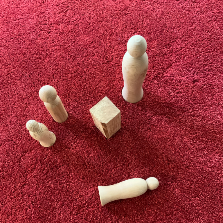 Fünf Holzfiguren und ein Würfel auf rotem Teppich angeordnet symbolisieren eine Gruppe Menschen mit einem Konflikt.